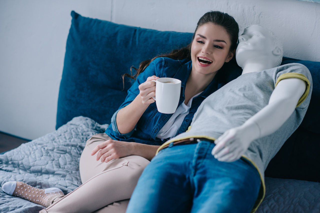 young woman with cup of coffee talking to mannequin while resting on bed, unrequited love concept